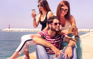 Group of travel friends having fun with mobile technology at ferry pier - Cheerful teenagers using phone sitting by ocean - University students on vacation for the spring break -Soft vintage filter