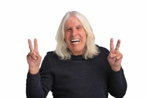 Senior man with shoulder length white hair giving peace signs with both hands and smiling.